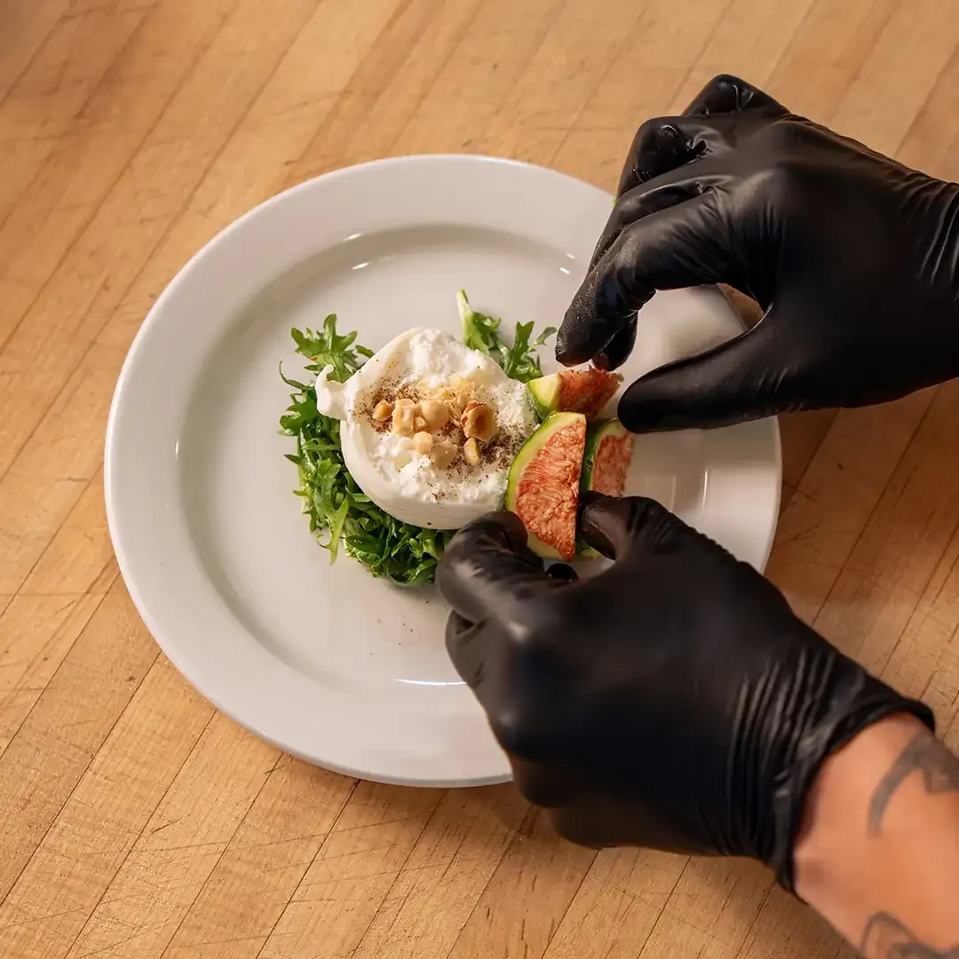 A chef in black gloves plating a Farm & Forage course with fresh greens